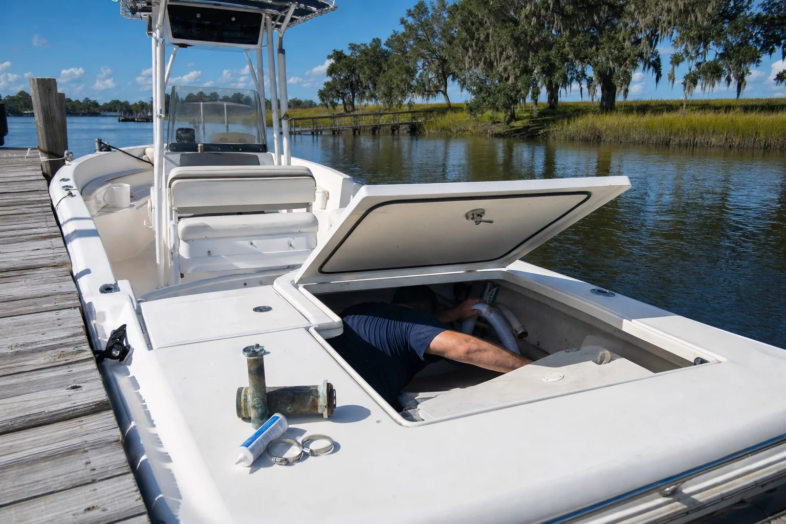 Technician inspecting engine compartment inside fiberglass boat at Wilmington dock with marsh and live oaks visible.